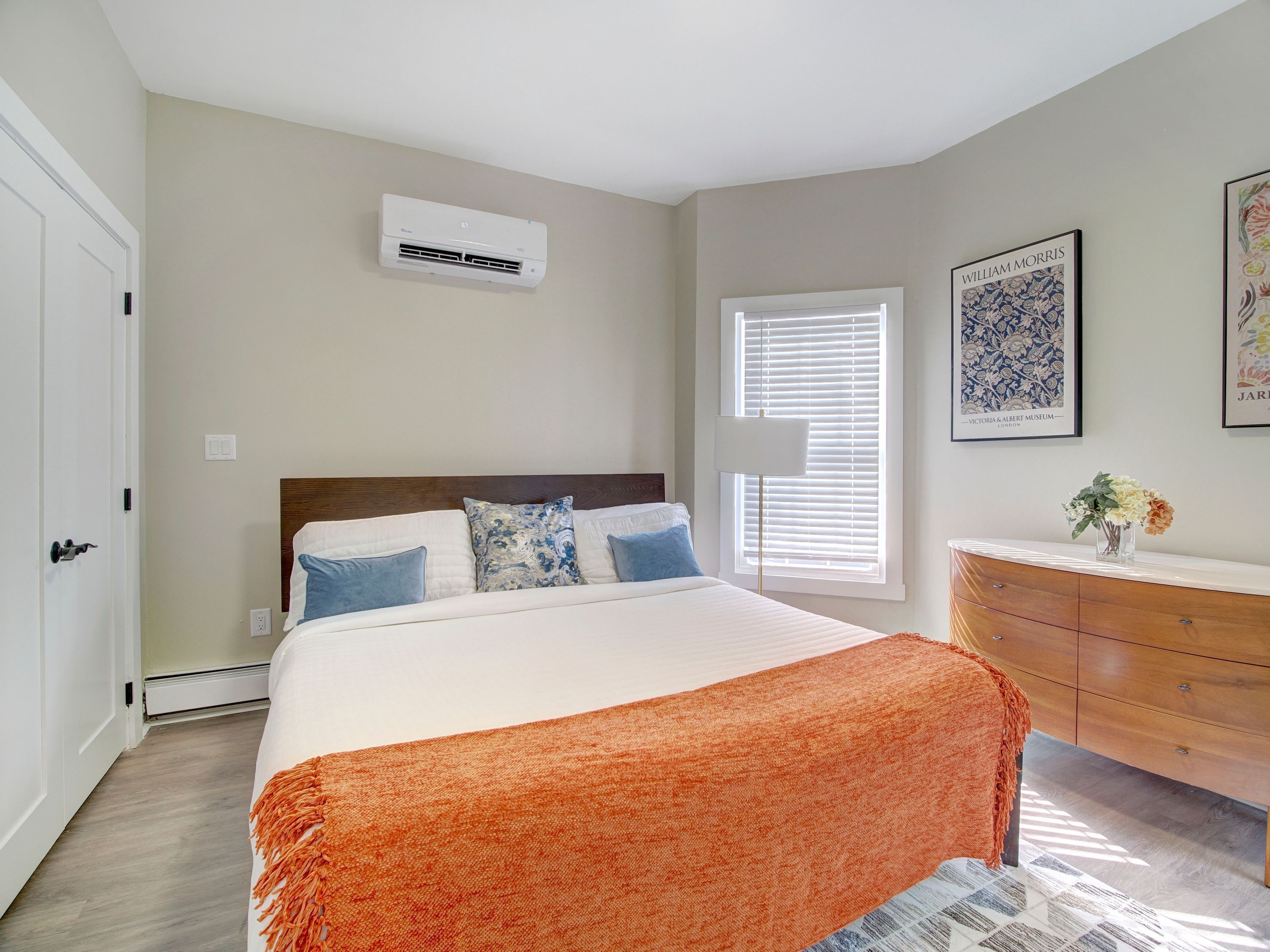 Une chambre à coucher comprenant un lit, une commode, un appareil de climatisation et deux fenêtres équipées de stores.