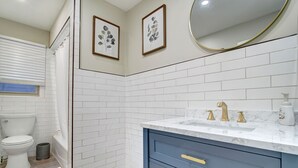 Un baño con paredes de azulejos blancos, un inodoro blanco, un tocador azul con pomos dorados y dos láminas enmarcadas con motivos botánicos.