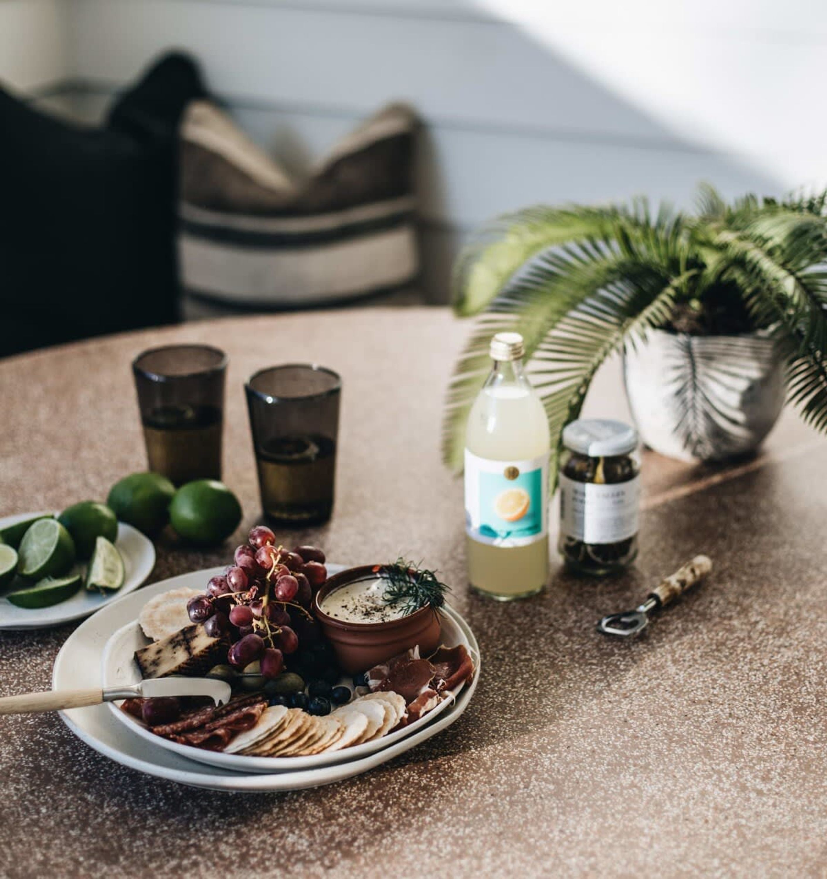 A table with a plate of sliced fruits, a bowl of dip, and a bottle of drink. There are also two glasses and a jar of jam.