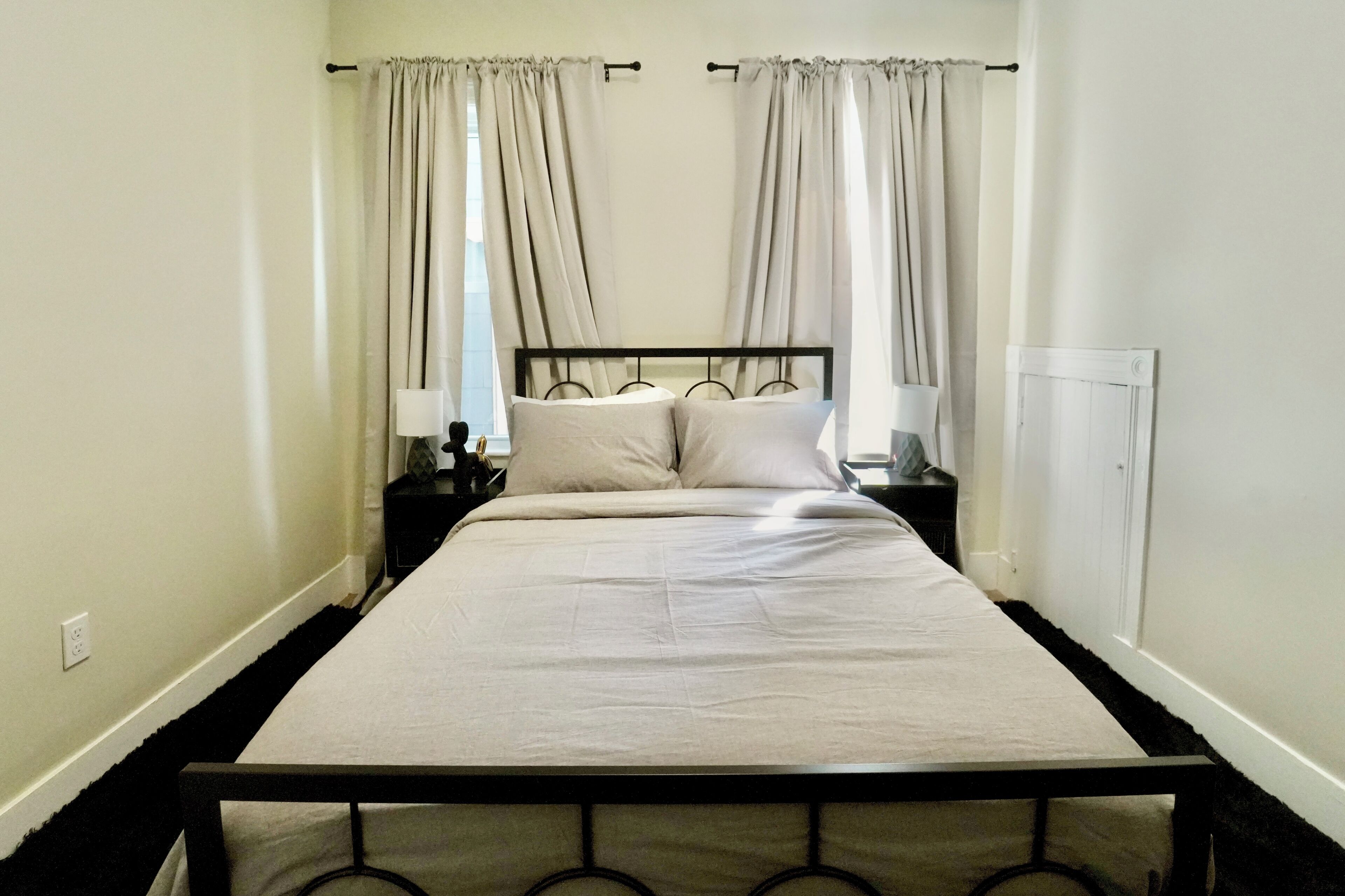 Una cama bien tendida con estructura de metal, dos mesitas de noche con lámparas y una ventana con cortinas.