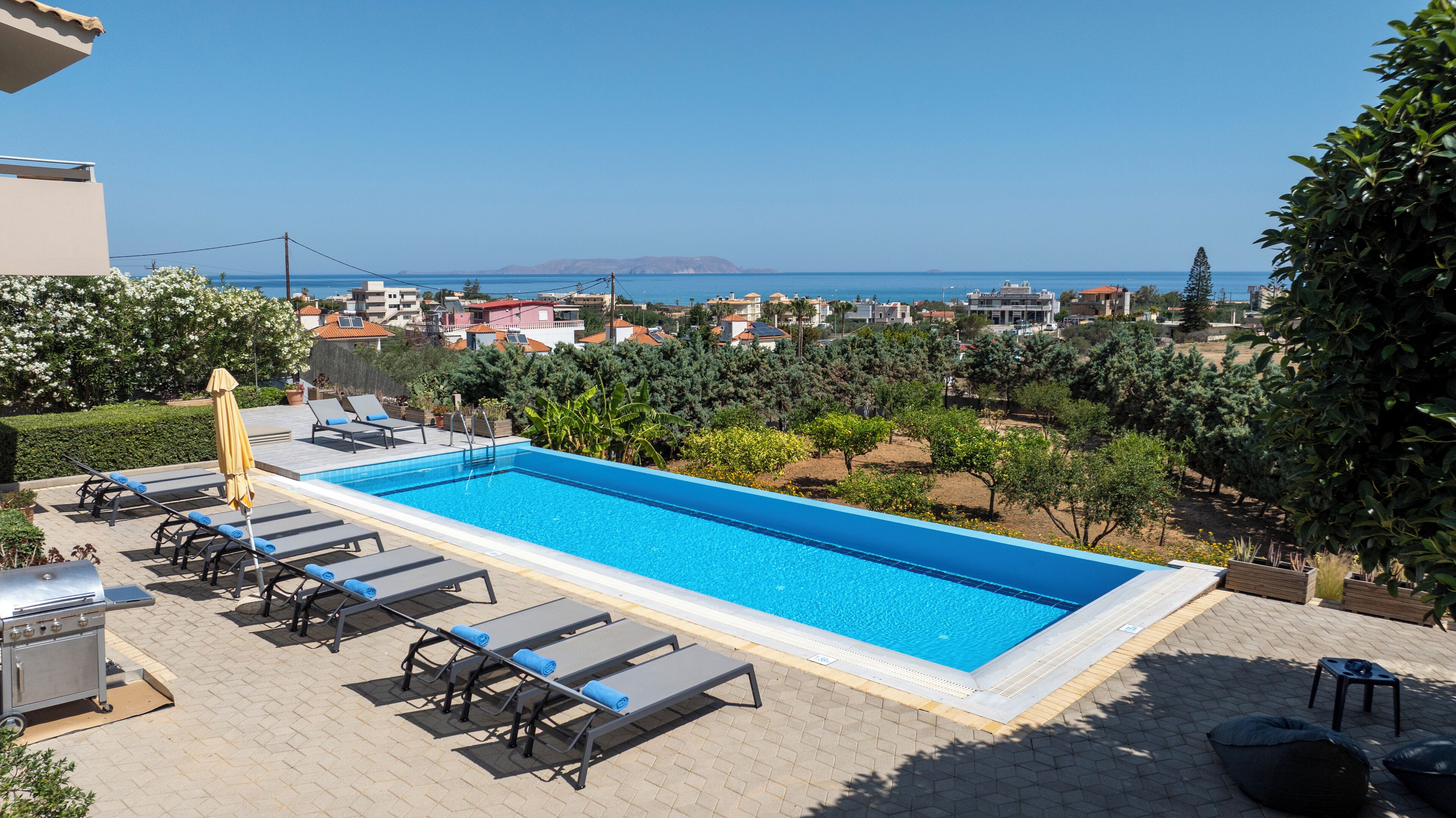 Une piscine rectangulaire à l’eau d’un bleu limpide, entourée de chaises longues et d’un barbecue, donnant sur un quartier résidentiel avec des maisons et offrant une vue sur la mer.