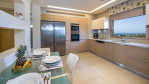 Una cocina moderna con refrigerador, horno y lavavajillas integrados. Hay una mesa de comedor dispuesta con platos, vasos y un frutero. Una ventana ofrece vistas al mar.