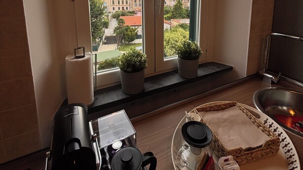 A kitchen with a coffee maker, a kettle, and a tray with cups and a plate. There are two windows with a view of buildings and trees. - Cappuccini's B&B (Livorno)