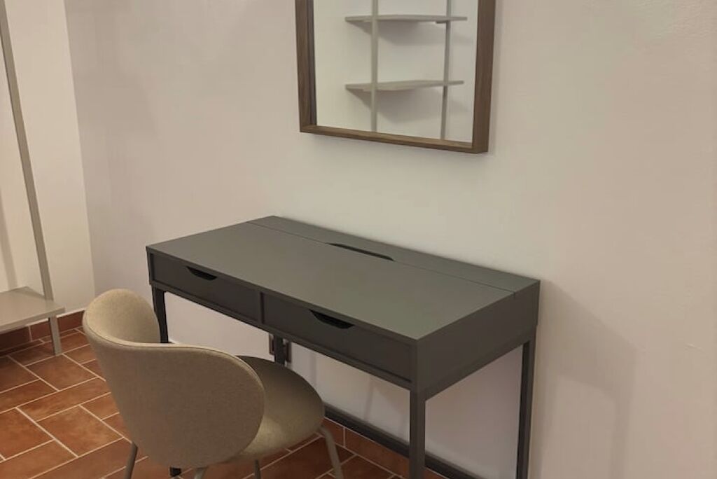 Une petite pièce moderne avec un bureau gris et une chaise, un mur blanc et une étagère avec un miroir.