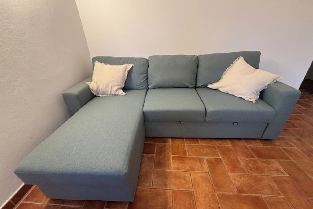 A light blue sectional sofa with two white pillows against a white wall and brown tiled floor.