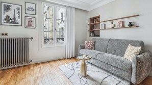 Un salón con un sofá gris, una pequeña mesa redonda, y un radiador. Hay cuadros enmarcados en la pared, una ventana con cortinas, y suelo de madera.