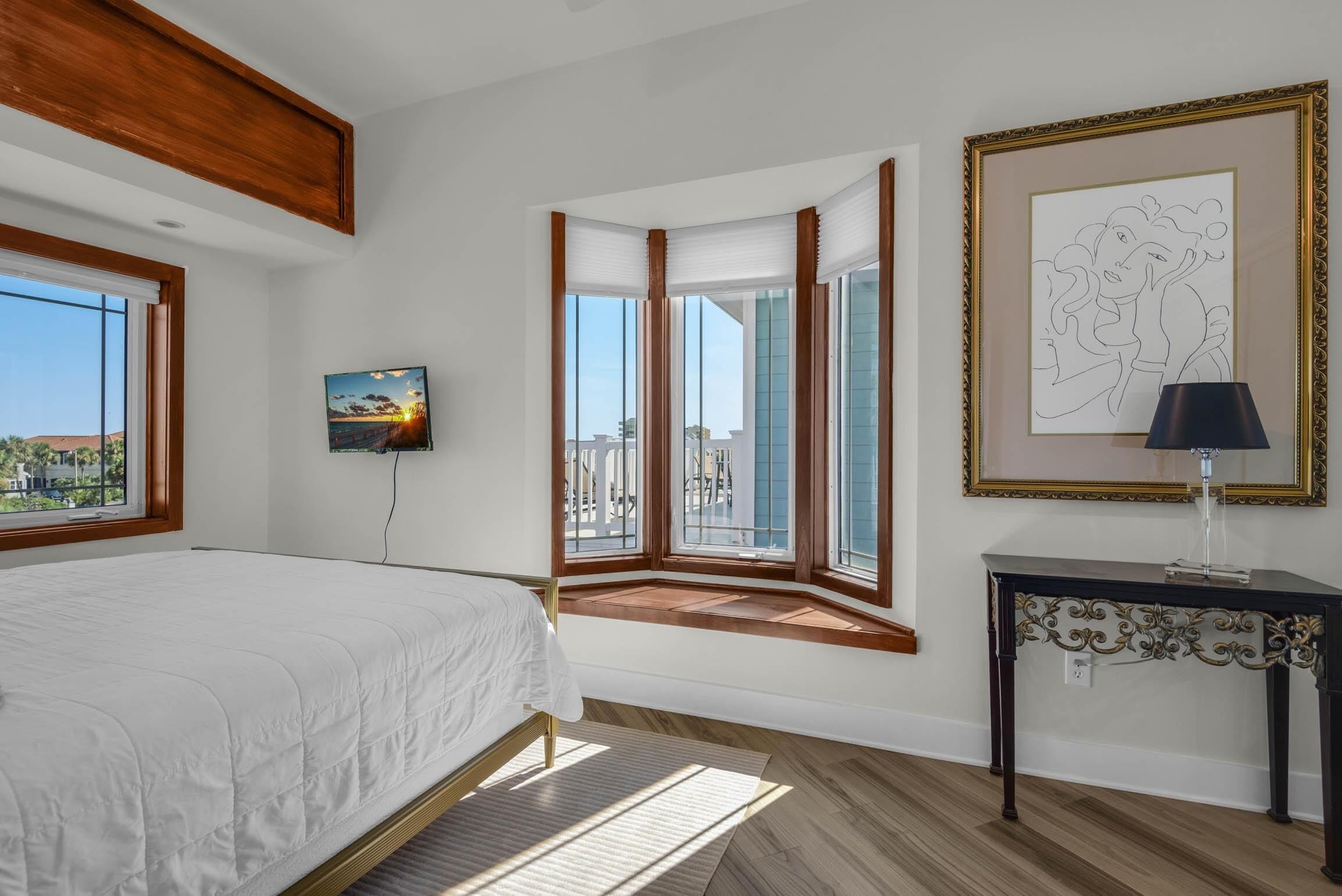 A bedroom with a large bed, a view through a window, a framed artwork, and a nightstand.