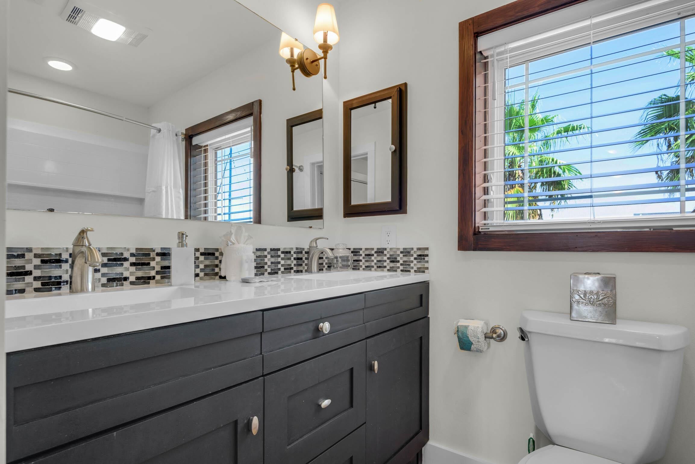Une salle de bain avec un lavabo blanc, des meubles sombres, des toilettes et une fenêtre avec des stores.