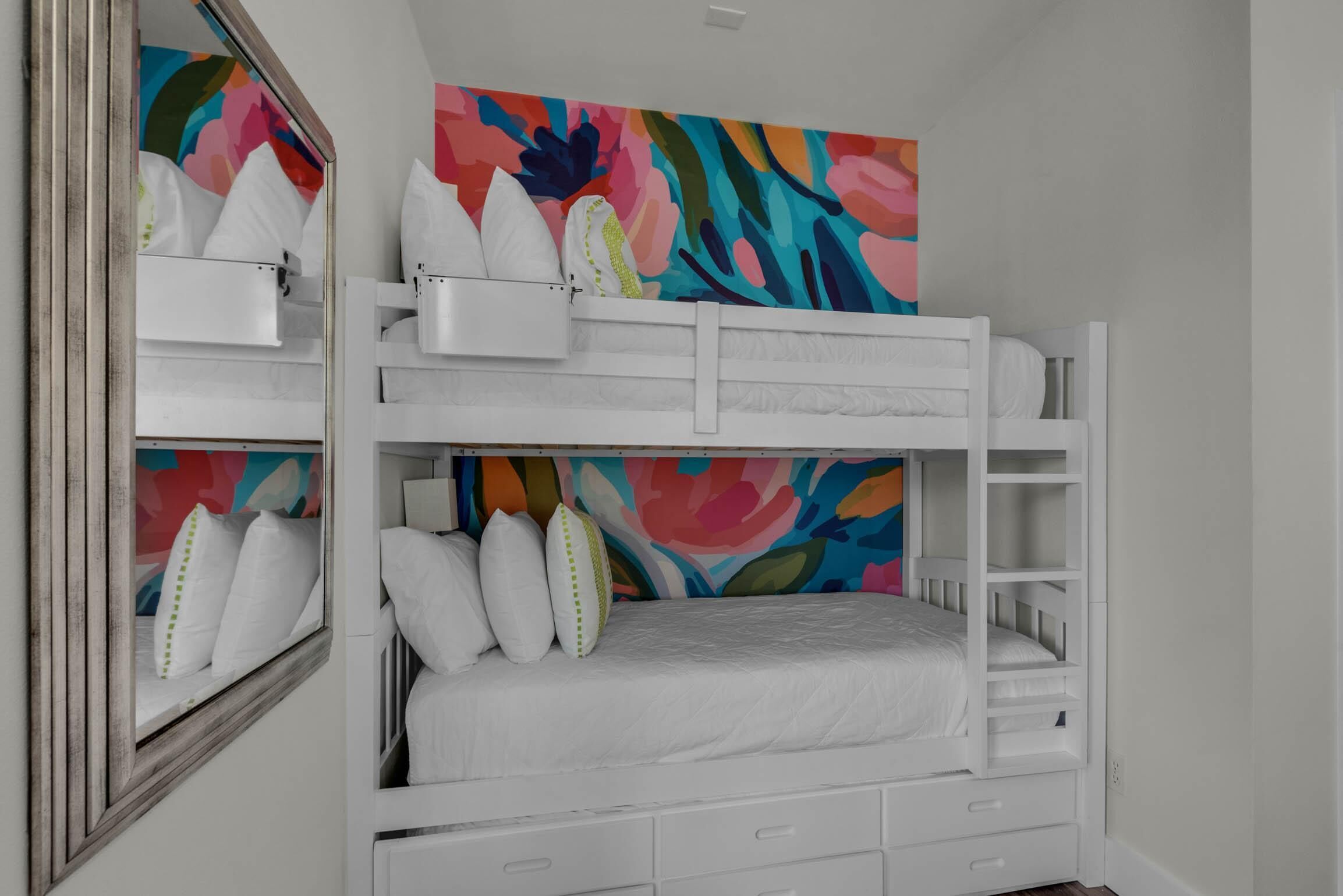 Una cama litera con ropa de cama y almohadas blancas, un cuadro abstracto colorido en la pared y un espejo a la izquierda.