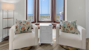 Ein Zimmer mit zwei weißen Sesseln, einem kleinen weißen Beistelltisch und einer Stehlampe. Große Fenster bieten Blick auf das Meer und die Stadt.