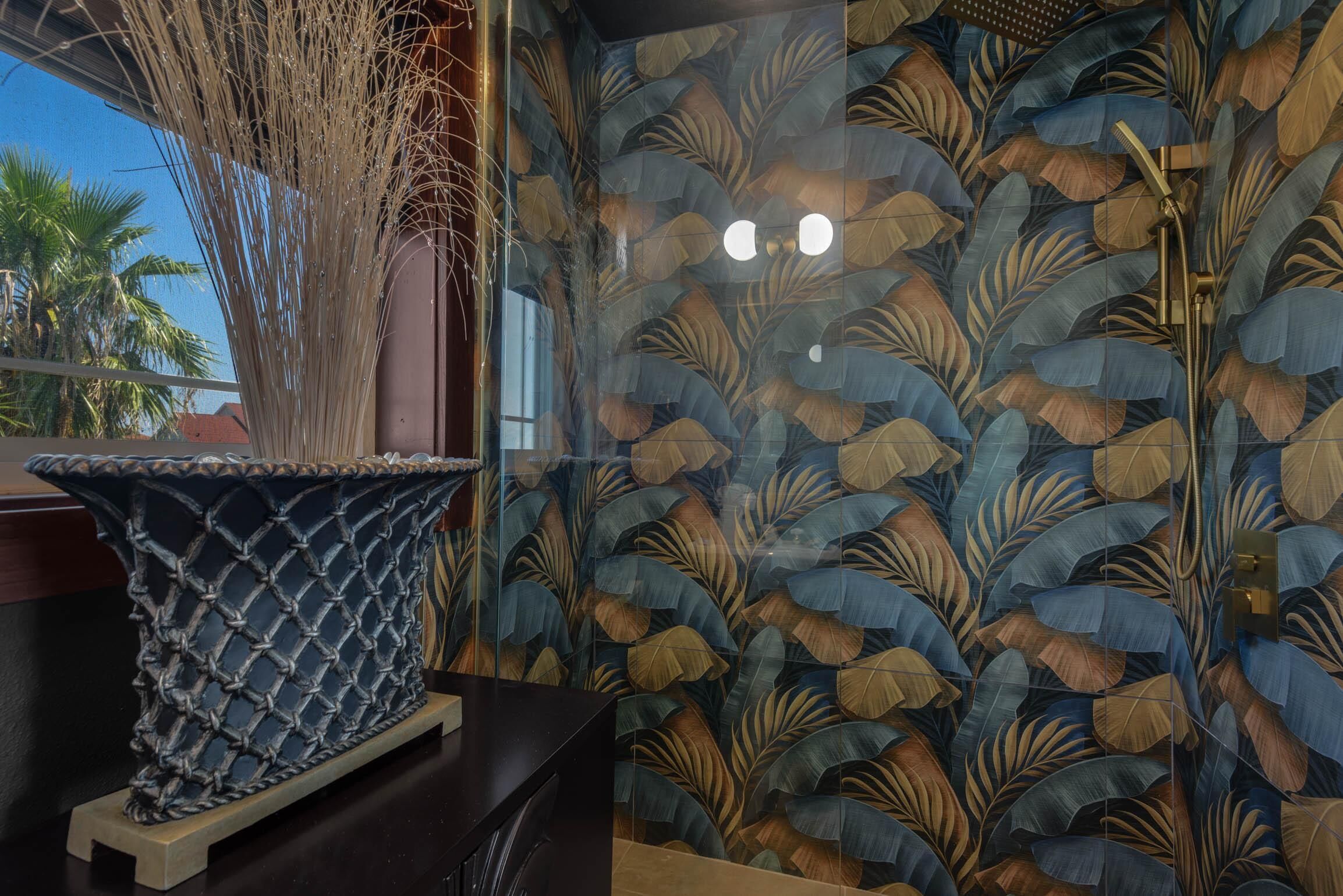 A bathroom with a decorative wall featuring leaf patterns, a shower area with a golden showerhead, and a decorative vase on a wooden stand.