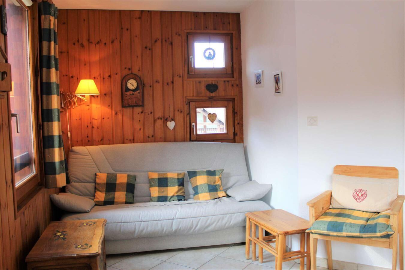 A cozy room with a sofa, a wooden chair, a small table, and a window with a clock and heart decorations.