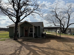 A small, single-story building with a porch and a covered area, surrounded by trees and a grassy field. - Private Furnished Guest House (Fort Worth)