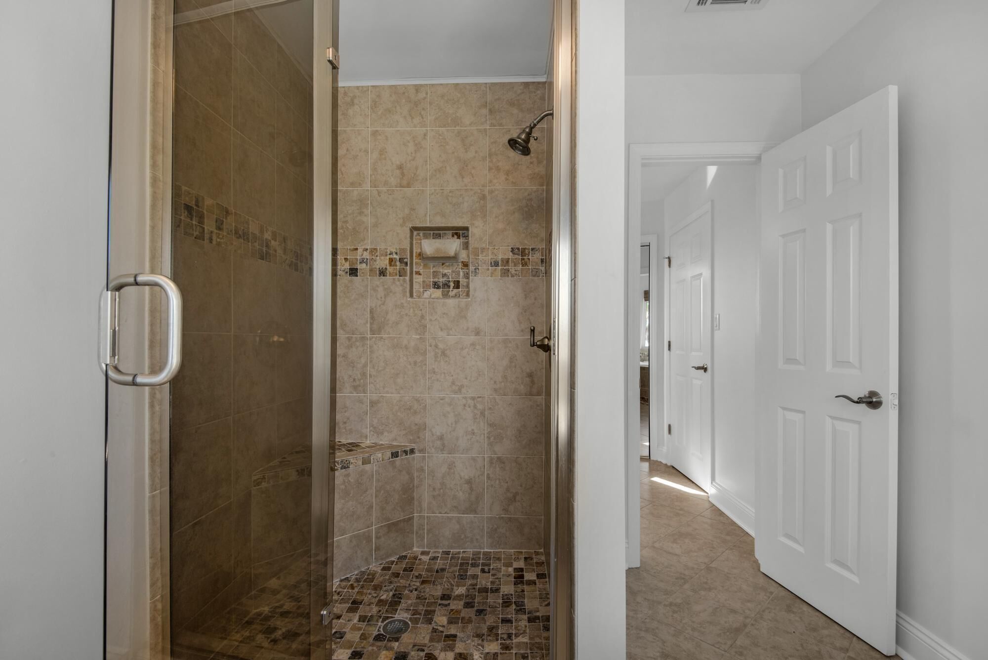 Une douche à l’italienne avec une porte en verre, des carreaux de couleur beige et une petite étagère carrelée.