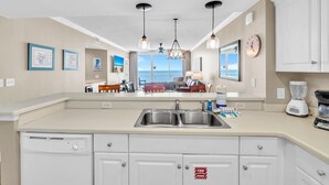 A modern kitchen with a central island, stainless steel sink, and a built-in dishwasher. There are pendant lights, a large window with a view, and a clock on the wall. - Majestic Beach Towers 2-1608 (Panama City Beach)