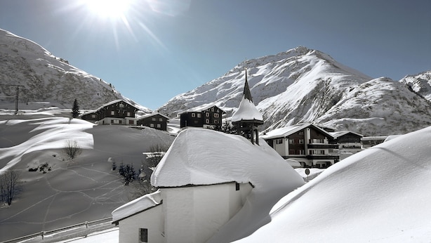 A snow-covered mountain village with chalet-style buildings, a church with a steeple, and a clear sky. - La Cruna Hotel-Restaurant (Tujetsch)