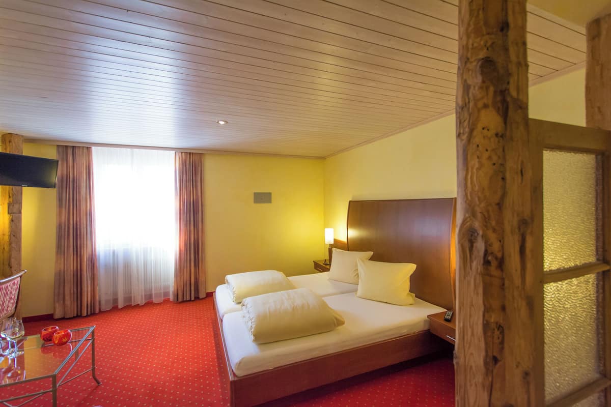 A hotel room with a wooden ceiling, a bed with white linens, a small table with a chair, and a TV mounted on the wall.