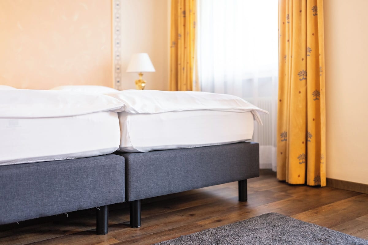A neatly made bed with white linens, a grey upholstered bench, and a small lamp on a bedside table.