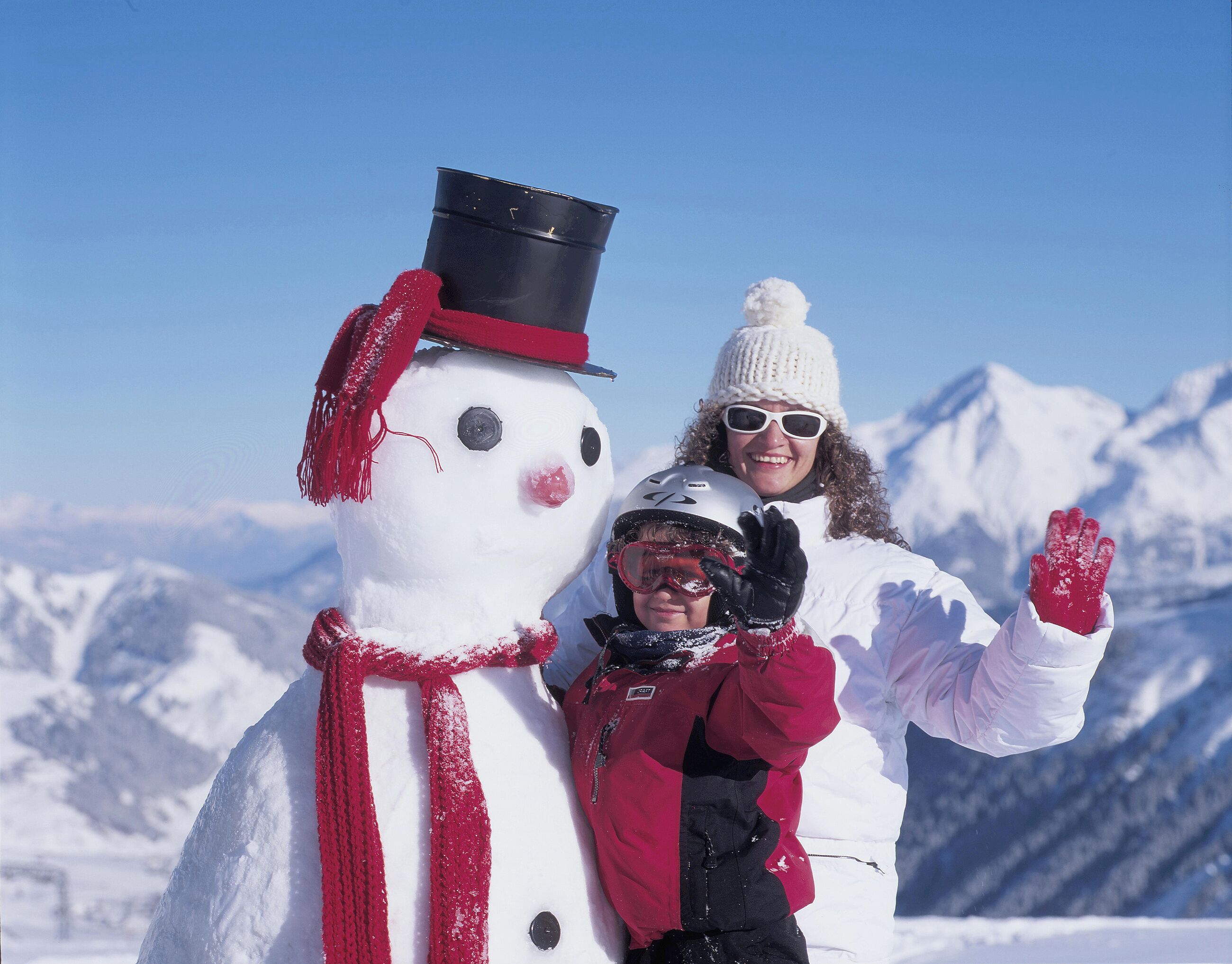 Ein Schneemann mit schwarzem Zylinder und rotem Schal, eine Person in einer weißen Jacke und ein Kind in roter Jacke mit Helm, stehen in einer verschneiten Landschaft mit Bergen im Hintergrund.