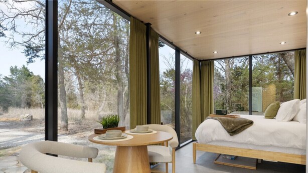 Une chambre moderne avec un grand lit, une table ronde en bois et une vue sur les arbres à travers de grandes fenêtres.