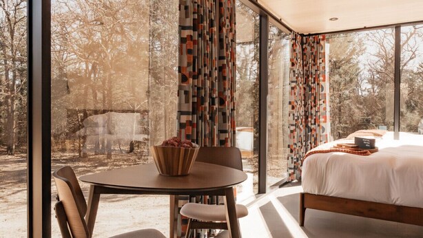 Un dormitorio moderno con una cama grande, una mesa de comedor con sillas y vistas a árboles a través de amplias ventanas.