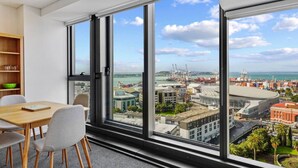 A modern room with large windows, a wooden table, and a view of a cityscape and port. - Modern Haven w Sea and City Views nr Spark Arena (Auckland CBD)