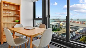 A modern dining area with a wooden table and chairs, a bookshelf with wine glasses, and a large window offering a view of a port and buildings. - Modern Haven w Sea and City Views nr Spark Arena (Auckland CBD)