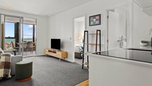 A modern living room with a TV, a wooden cabinet, a small green ottoman, and a view of the outside. - Modern Haven w Sea and City Views nr Spark Arena (Auckland CBD)