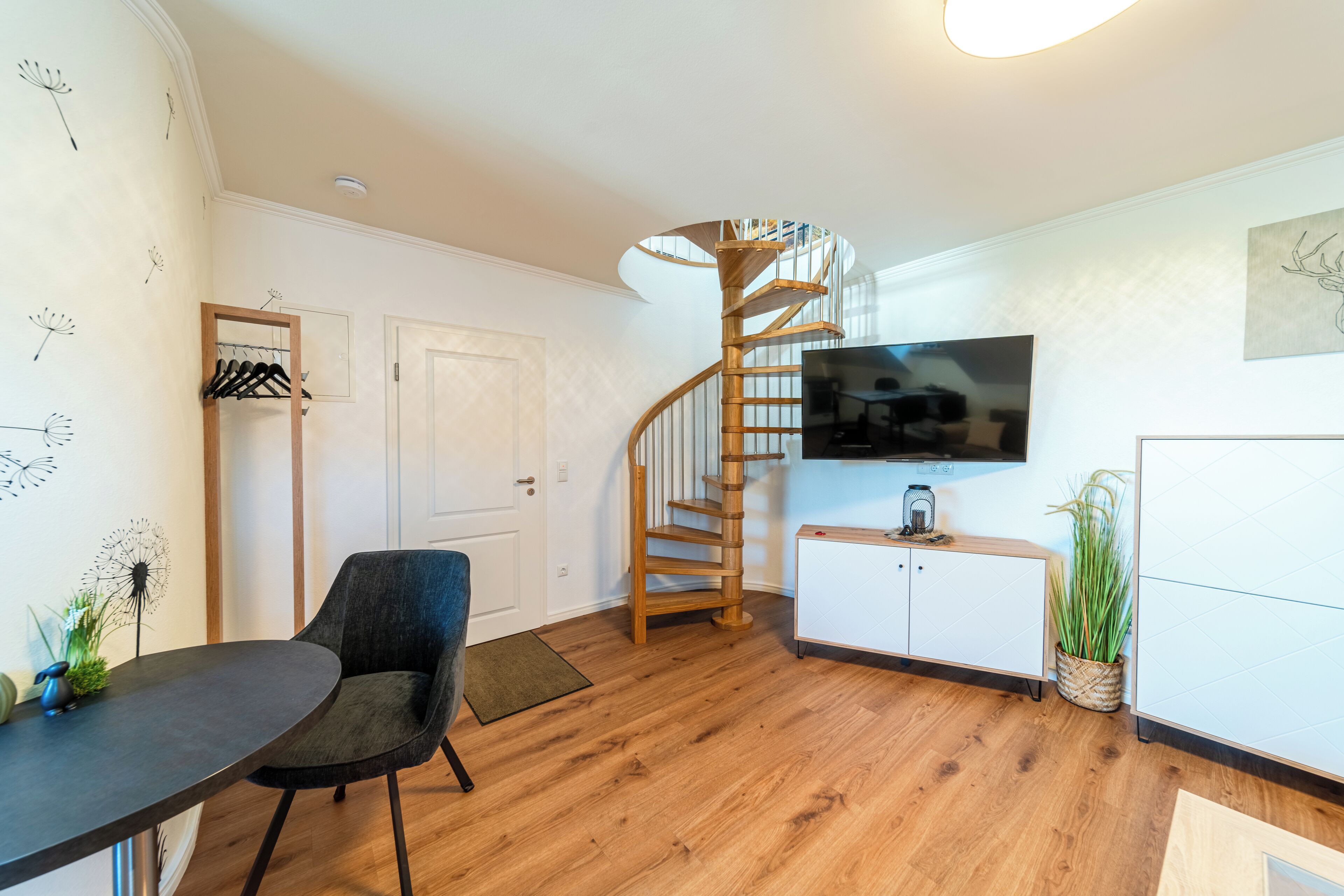 A modern living room with a wooden spiral staircase, a flat-screen TV, a dining table, and a seating area.