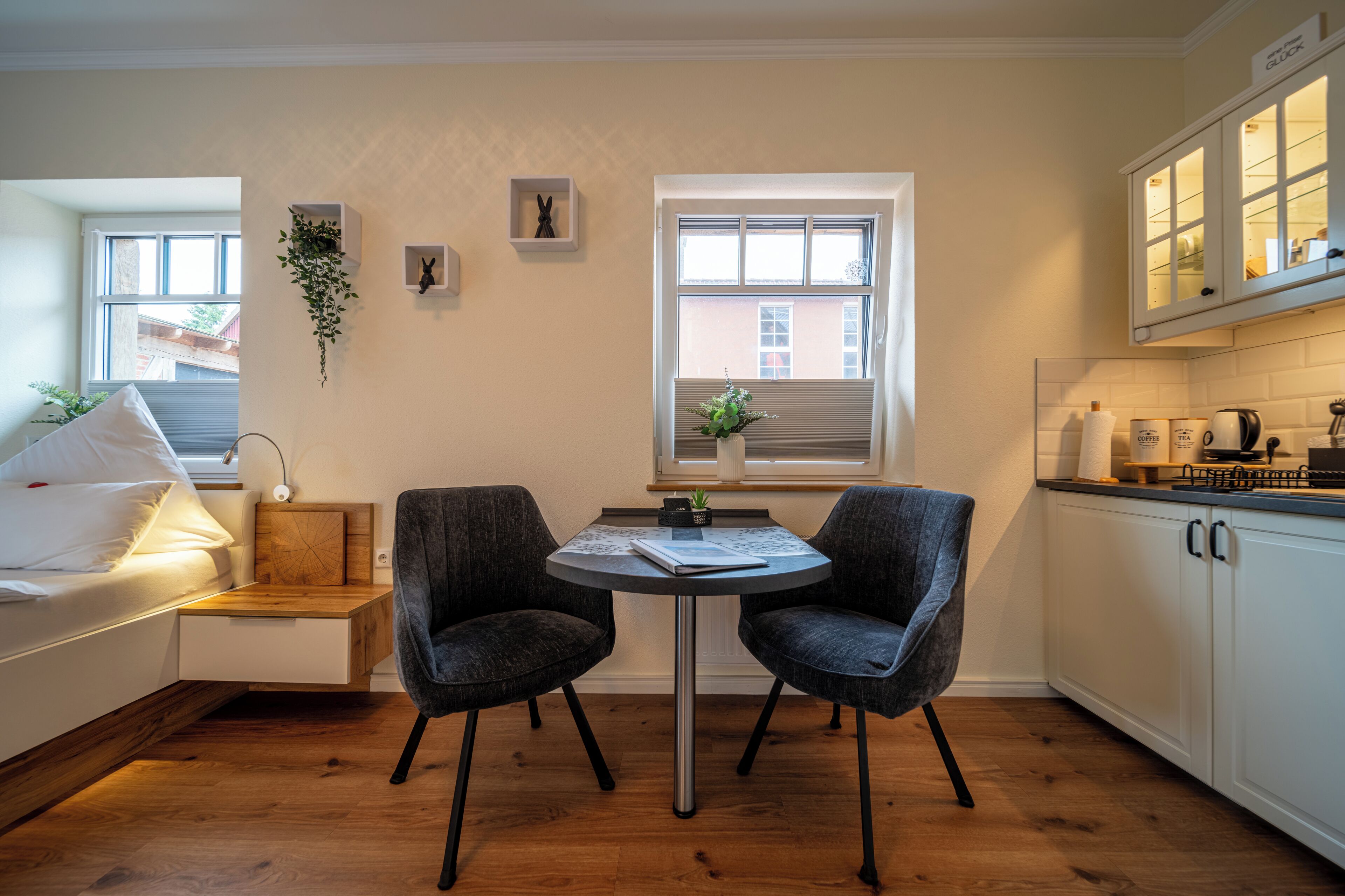 Une chambre moderne avec un lit, une table de chevet et un petit bureau avec deux chaises. Il y a une fenêtre avec des stores, une plante et une vue sur un bâtiment à l’extérieur.