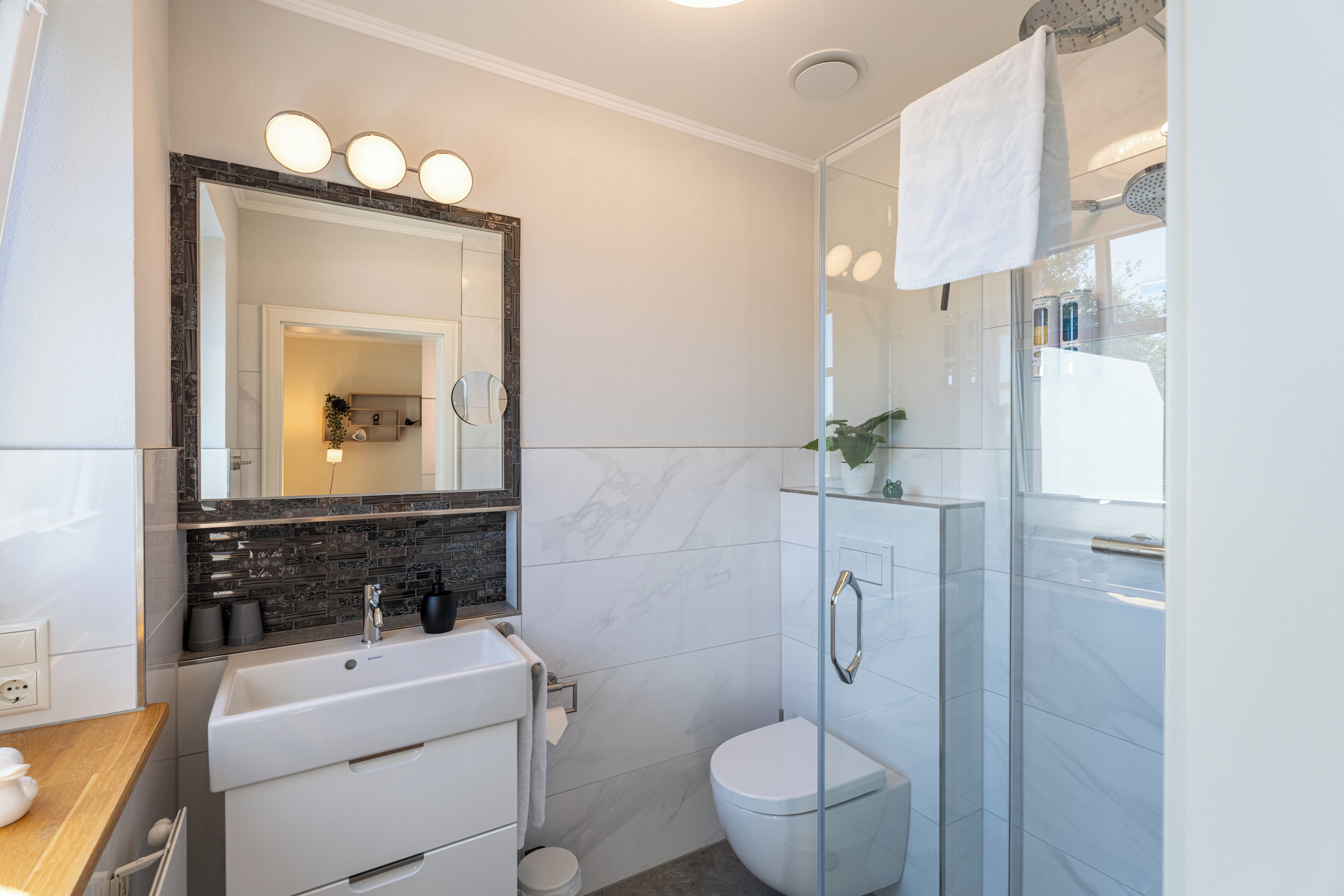 A modern bathroom with a white sink, a glass shower enclosure, and a large mirror with lights.
