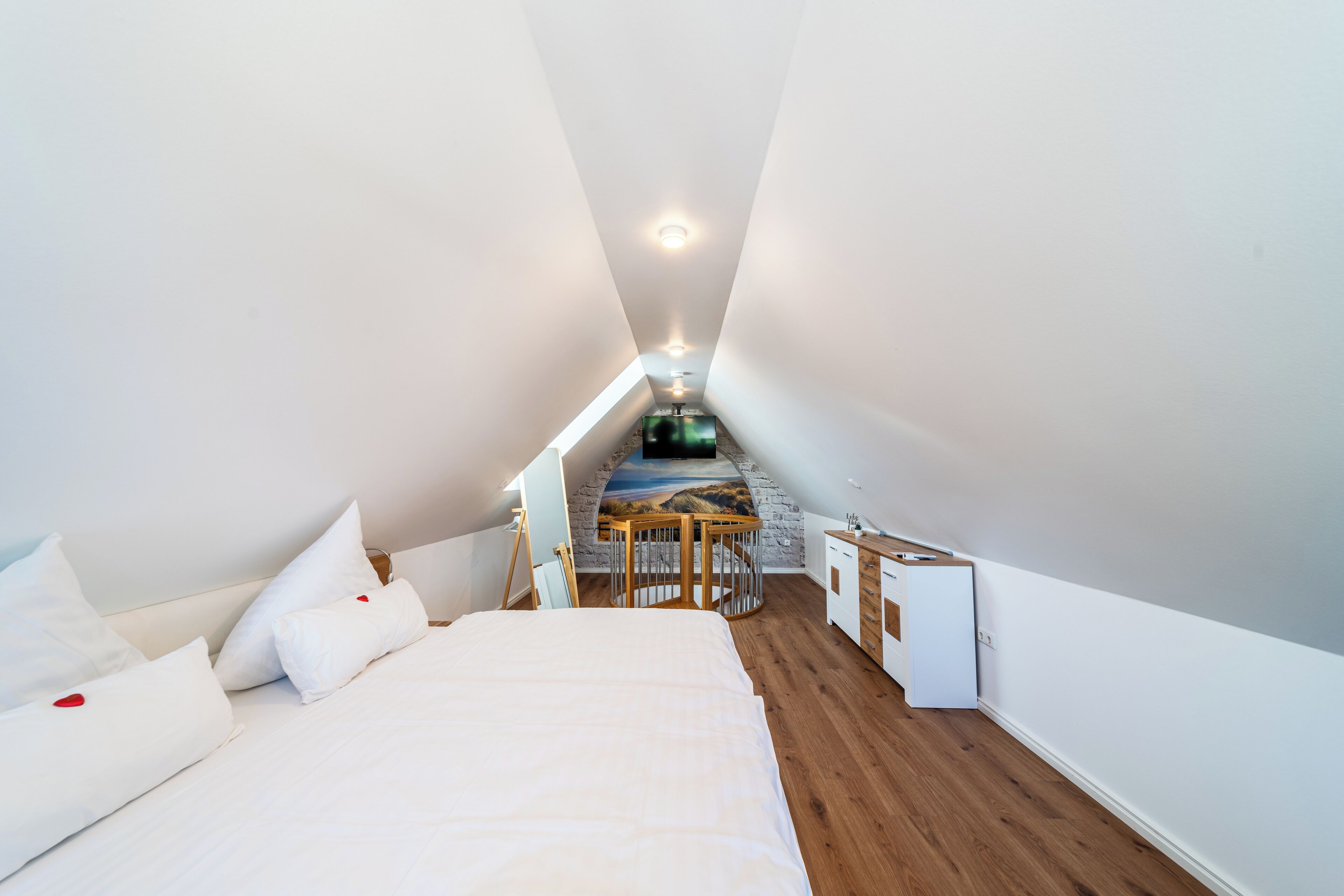 Una habitación estrecha y bien iluminada con una cama, una mesita y un televisor.