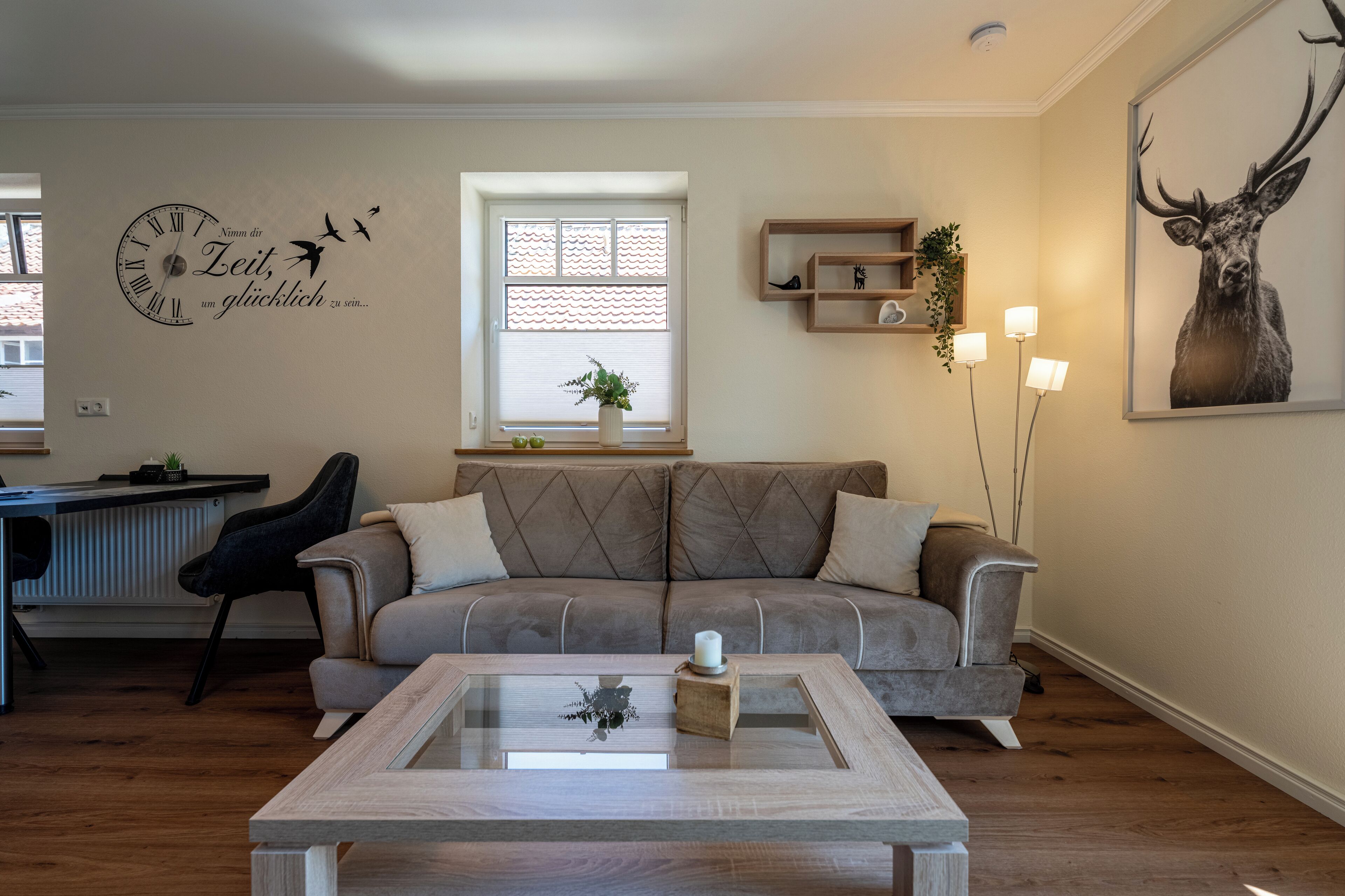 Una sala de estar con un sofá gris, una mesa de centro de vidrio, un reloj de pared, una ventana con persianas y un cuadro con una imagen de un ciervo.
