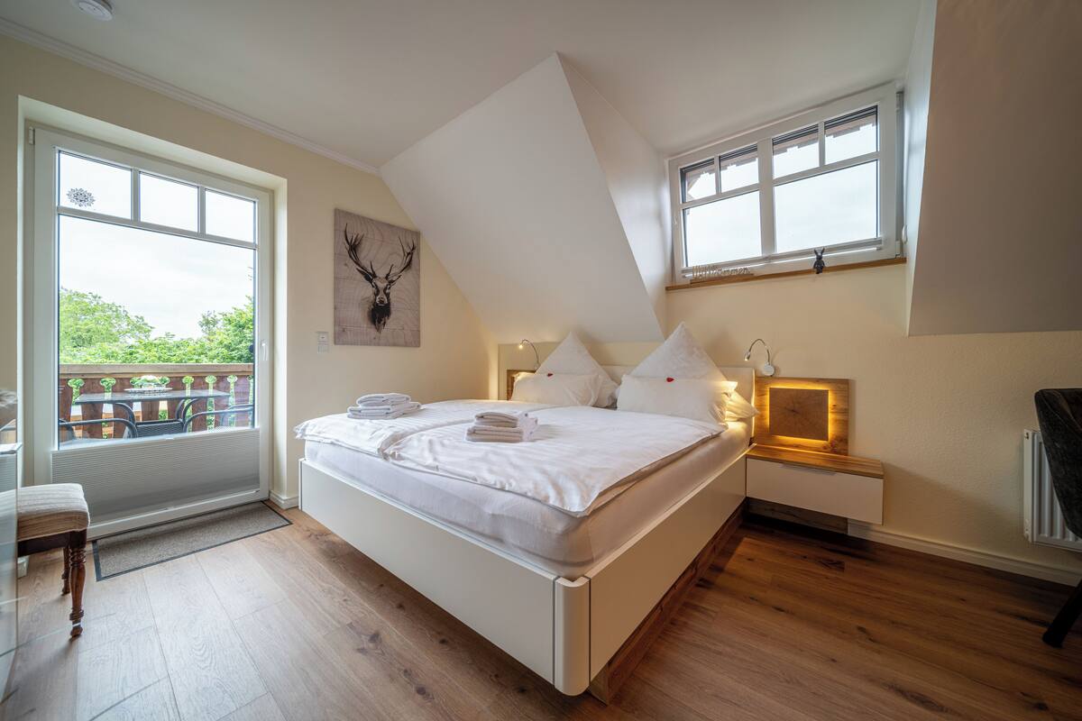 A bedroom with a large bed, a window, a wooden floor, and a view of greenery outside.