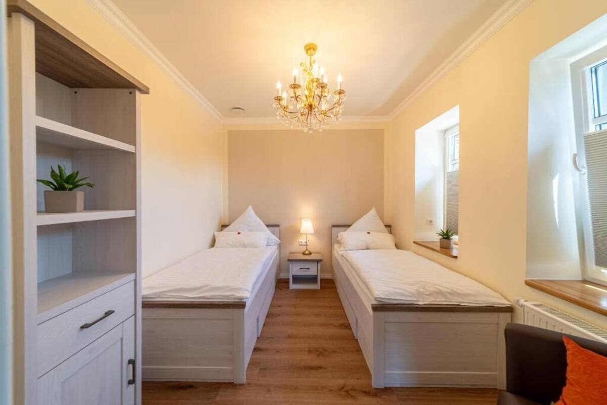 A bedroom with two beds, a chandelier, a nightstand, and a window with blinds.