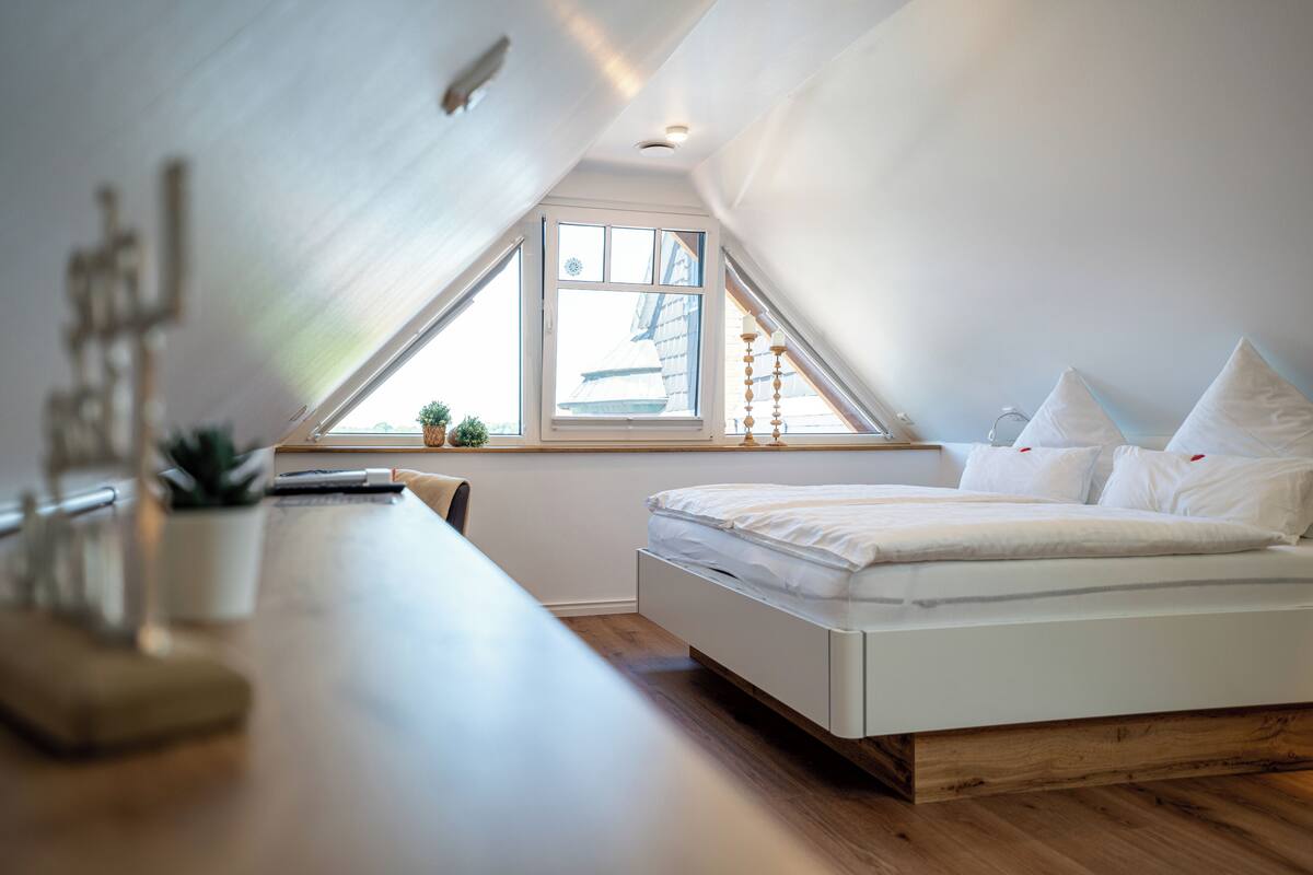 A bedroom with a sloped ceiling, a bed, a desk, and a window with a view of the outside.