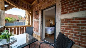 Een balkon met een glazen tafel en stoelen, een bakstenen muur en uitzicht op een tuin en gebouwen.