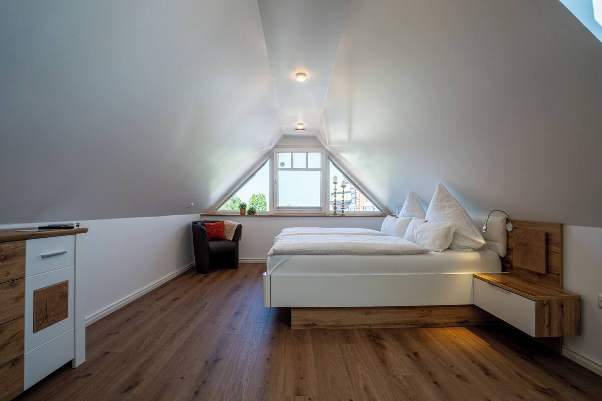 A bedroom with a sloped ceiling, a bed, a nightstand, a dresser, and a window with a view of greenery.