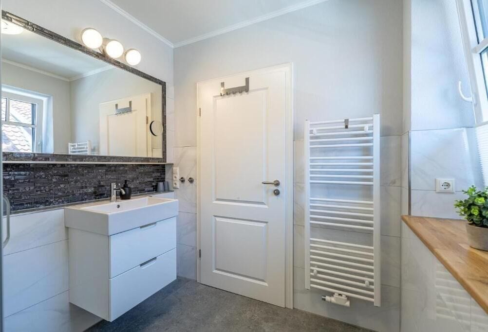 Un baño moderno con un tocador blanco, un espejo grande con luces, una puerta blanca y un perchero para toallas.