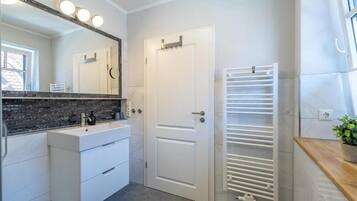 Un baño moderno con un tocador blanco, un espejo grande con luces, una puerta blanca y un perchero para toallas.