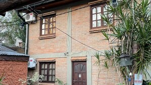 Un edificio rústico de ladrillo con una puerta de madera marcada con el número 3, un pequeño jardín con plantas en macetas y un banco de madera, y una lámpara colgante.