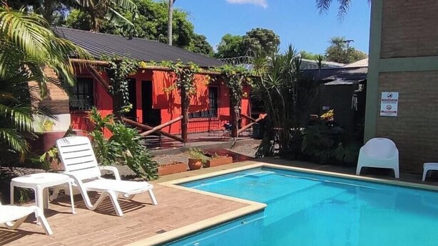 Un área de piscina con sillones blancos, una terraza de madera y un edificio rojo con un porche cubierto.