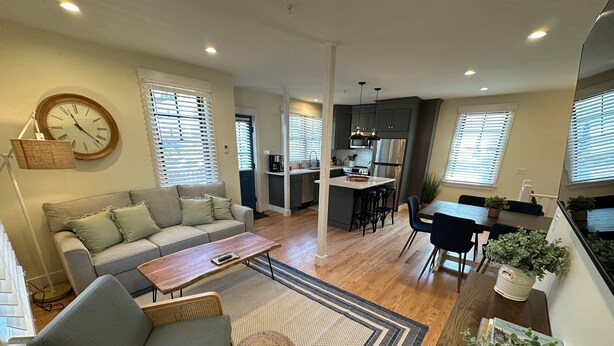 A modern living room with a sofa, coffee table, and ottoman, a dining area with a table and chairs, and a kitchen with a refrigerator and bar stools.