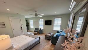 A bedroom with a bed, bedside table, and lamp. There is a living area with a sofa, coffee table, and TV. The room has wooden flooring and a ceiling fan.
