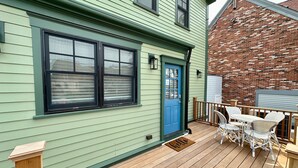A green house with a blue door and windows, a wooden deck with outdoor furniture, and a brick wall in the background.