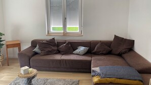 A living room with a brown sofa, a small wooden side table, a floor lamp, and a window with blinds. - Vacation apartment on the Danube cycle path (Untermarchtal)