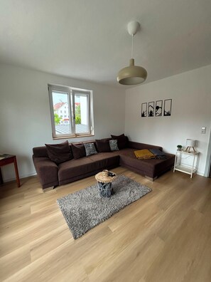 A modern living room with a sectional sofa, a small round side table, a floor lamp, and a large window with a view of buildings. - Vacation Apartment on the Danube Bike Path (Untermarchtal)
