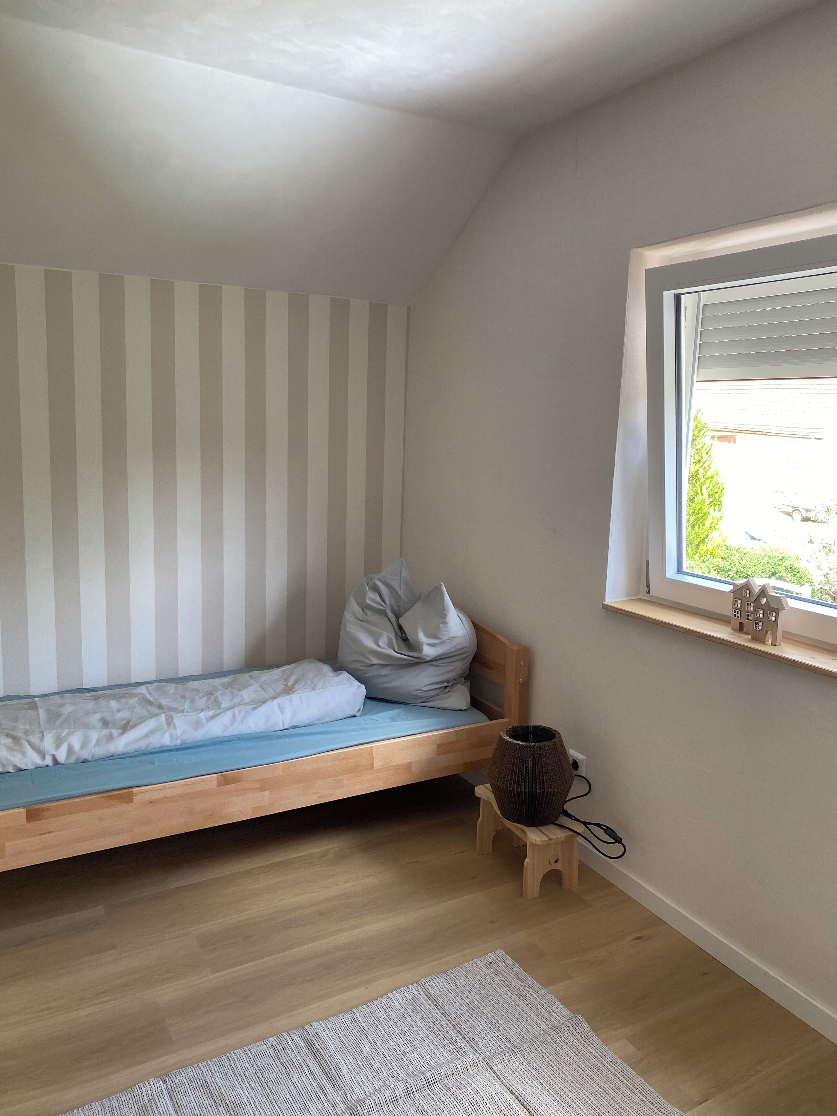 A small, single-bed room with a sloped ceiling, a window with blinds, a wooden bedside table, and a small rug on the floor.