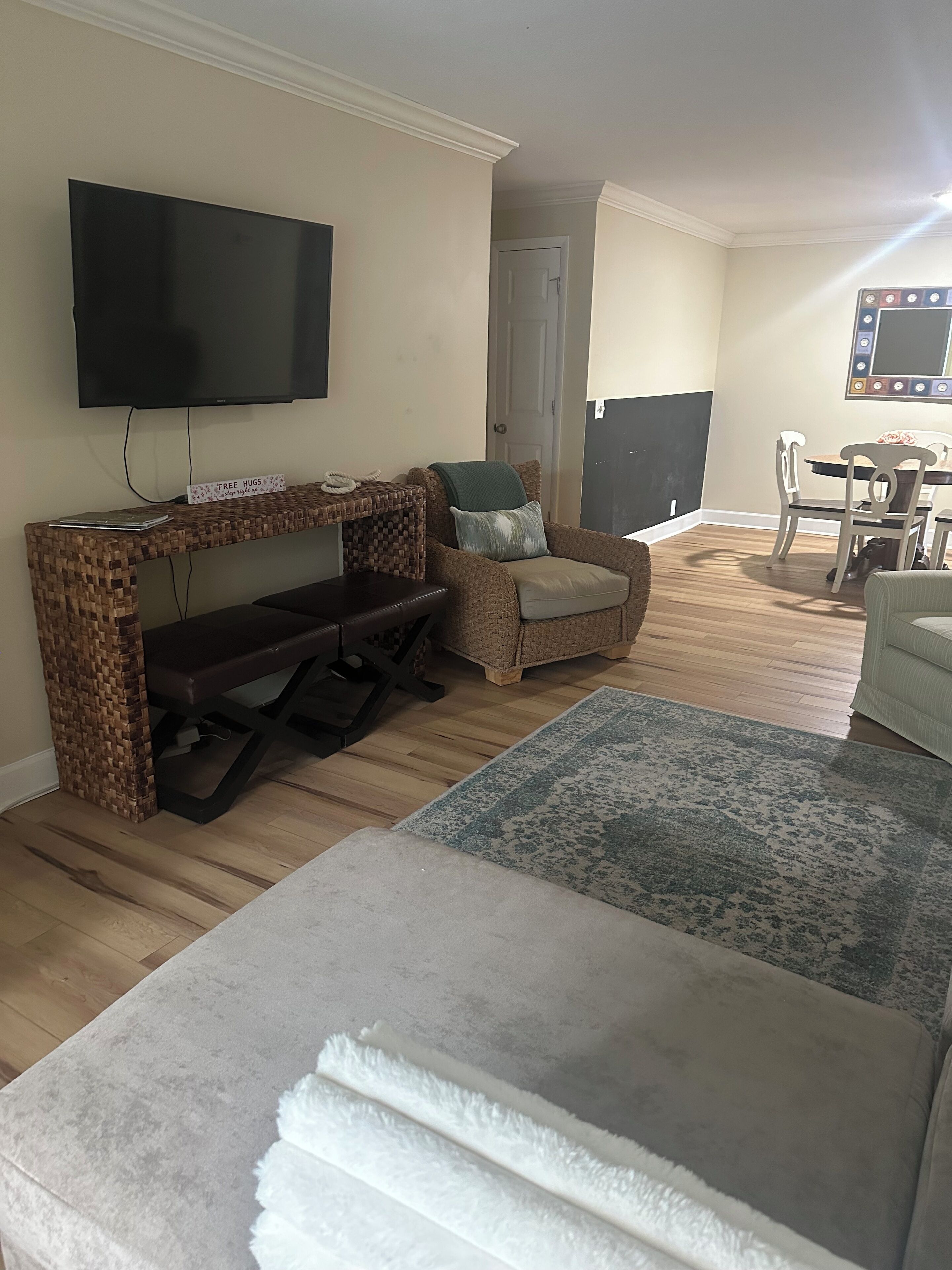 A living room with a flat-screen TV, a woven bench, a cushioned chair, a dining table with chairs, and a large rug.