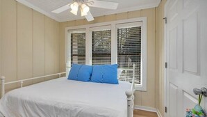 A bedroom with a white bed, blue pillows, a ceiling fan, and a window with blinds.